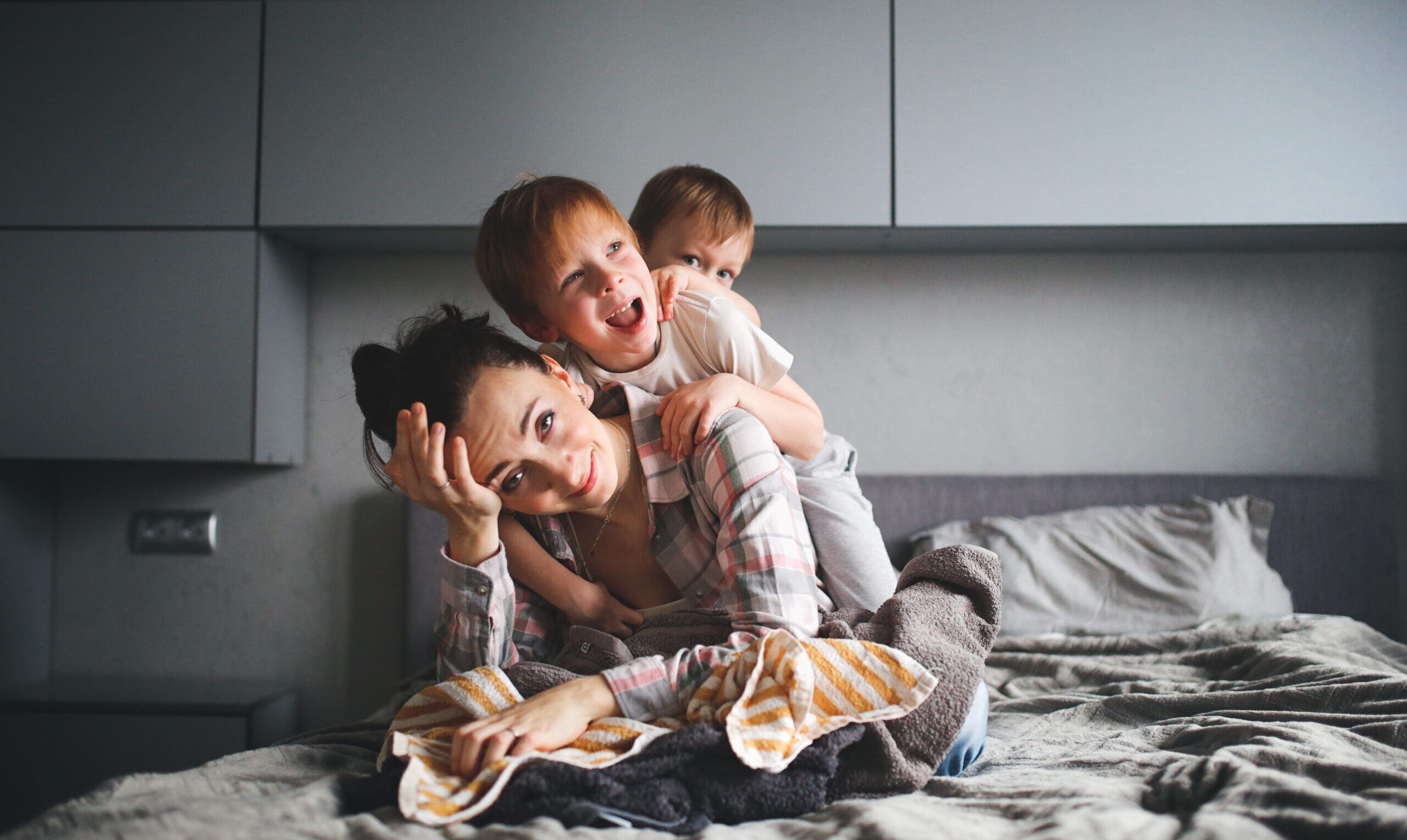 tired mother with two children, crazy children hang on mother in a gray real bedroom. Authentic lifestyle and motherhood difficulties.